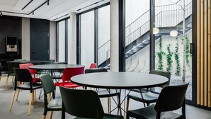 Moderni sisätila, jossa on pyöreitä pöytiä ja tuoleja, suuret ikkunat sekä portaikko, joka johtaa ulkoalueelle, jossa on viherkasveja.
