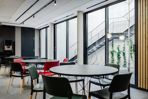 Un espacio interior moderno con mesas y sillas redondas, ventanas amplias y una escalera que conduce a un área exterior con vegetación.