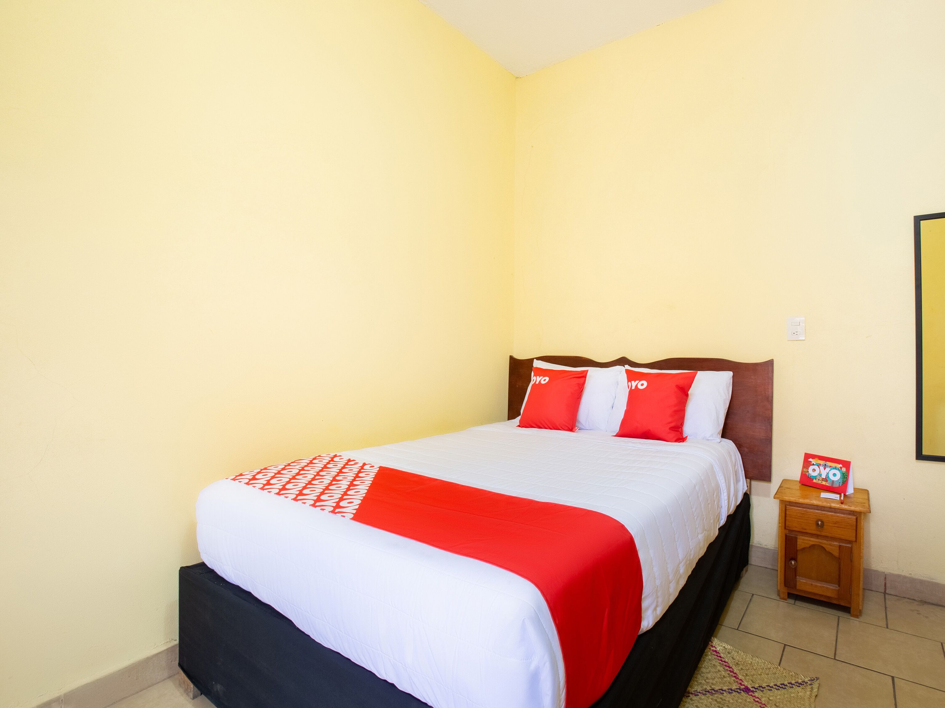 A neatly made bed with a red and white bedspread and pillows, a wooden nightstand with a book, and a plain yellow wall.