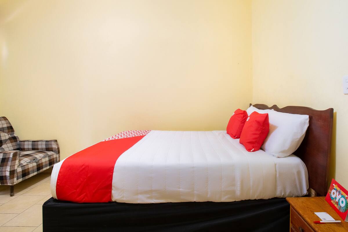 A neatly made bed with a red and white bedspread, a wooden headboard, and a plaid armchair.