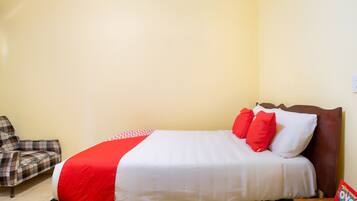 Un lit bien fait, recouvert d’une couvre-lit rouge et blanche, avec une tête de lit en bois et un fauteuil à motifs écossais.