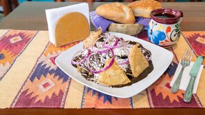 Un plato de enchiladas con guarniciones, una cesta de pan, un frasco de salsa y una servilleta con cubiertos sobre un mantel estampado.
