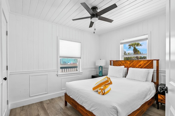 Un dormitorio con cama de madera, paredes blancas, ventilador de techo, dos ventanas y vista a palmeras.