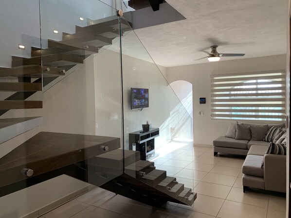 A modern living room with a glass staircase, a ceiling fan, a flat-screen TV, and a sofa.