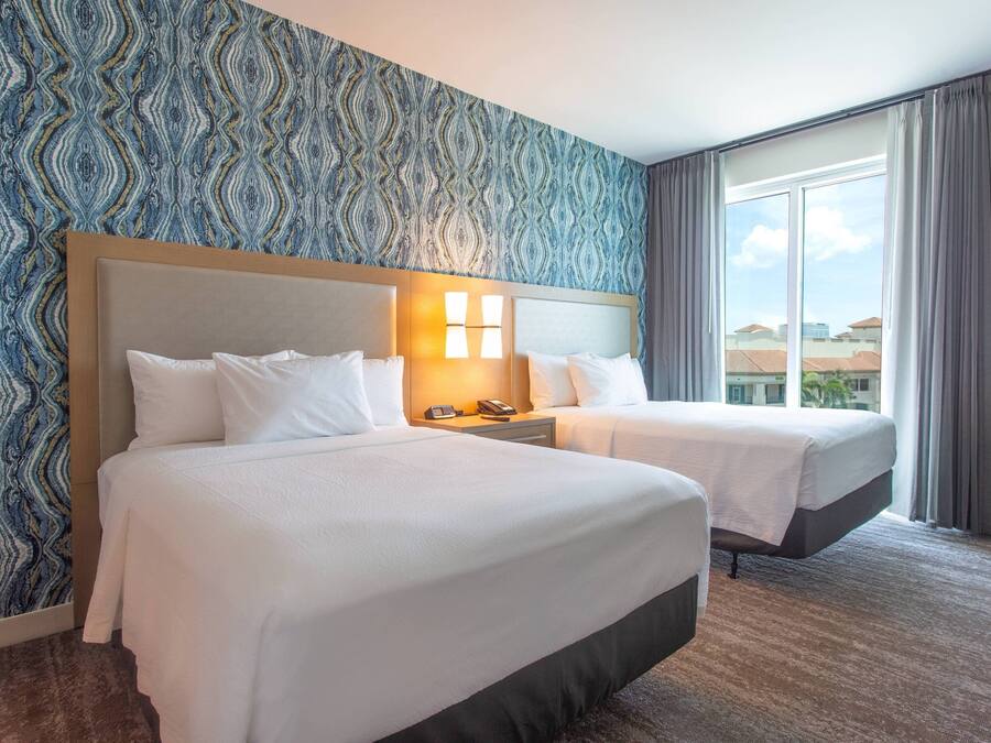 Habitación de hotel con dos camas, papel tapiz estampado, un ventanal con cortinas y vista a edificios.