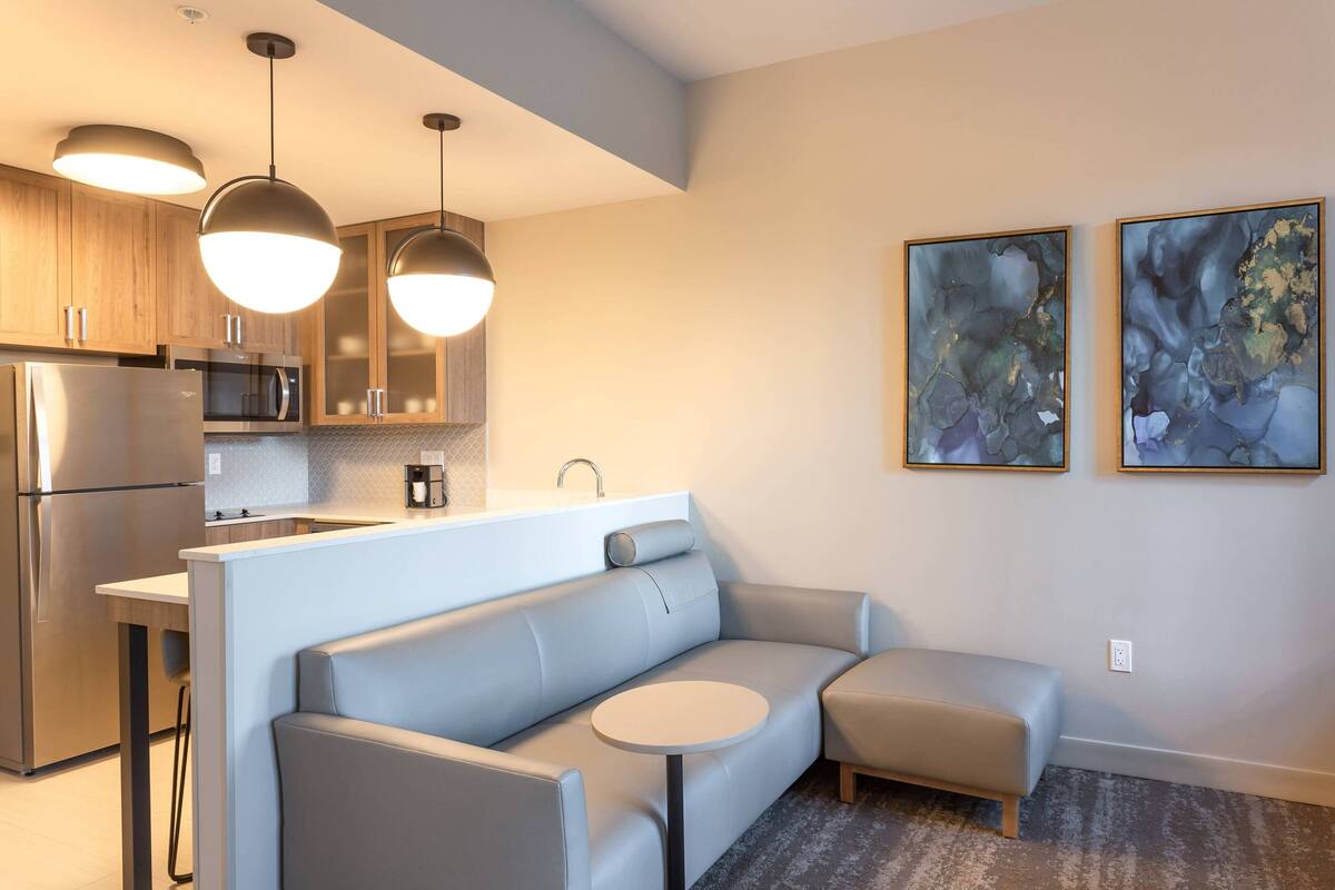 A modern kitchen with stainless steel appliances, a built-in oven, and a bar area with a light-colored countertop. There is a gray sofa and a small round table in the living area.