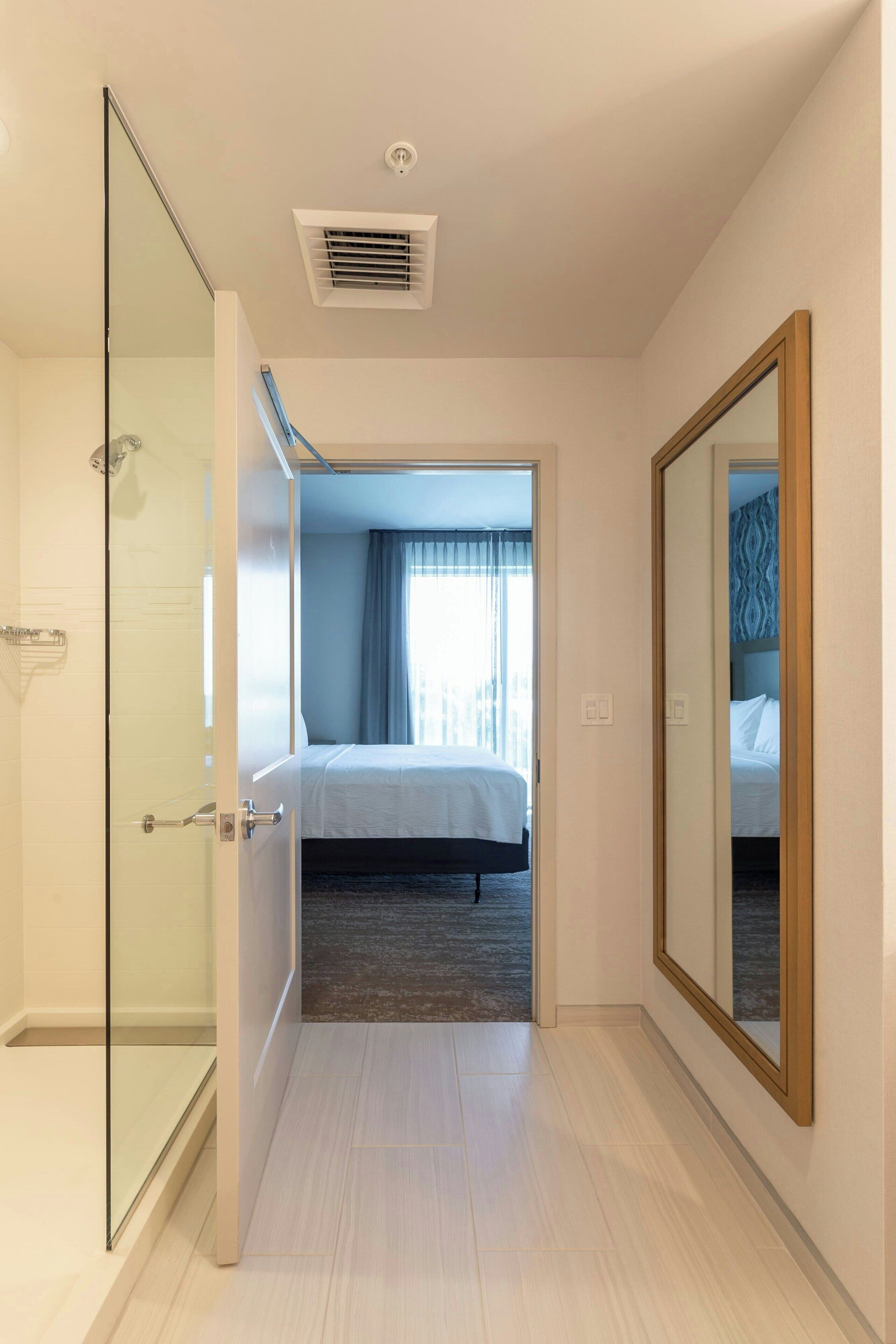 Une chambre d’hôtel dotée d’une douche en verre, d’un lit avec une tête de lit sombre et d’un grand miroir au mur.