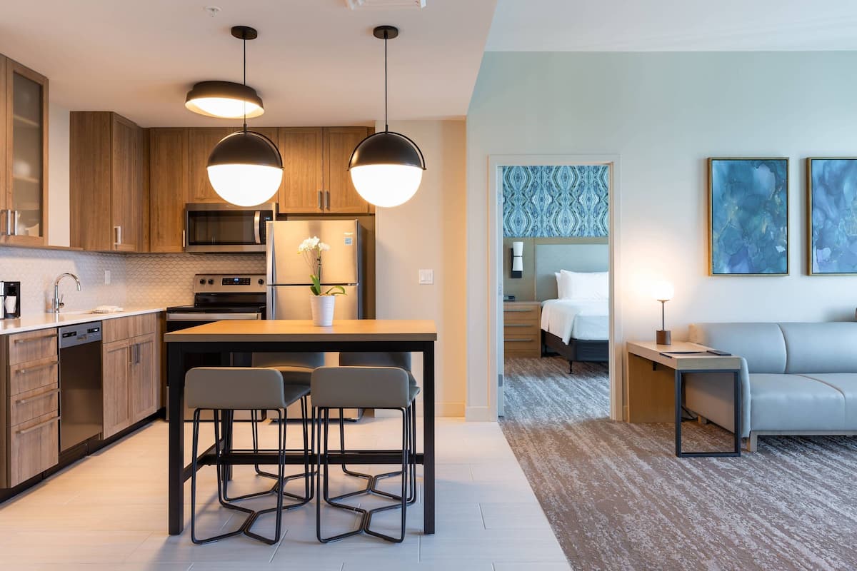 A modern kitchen with wooden cabinets, a dining area with two chairs, a small table, and a refrigerator. There is a microwave, an oven, and a coffee machine. The living area has a sofa and a small table with a lamp.
