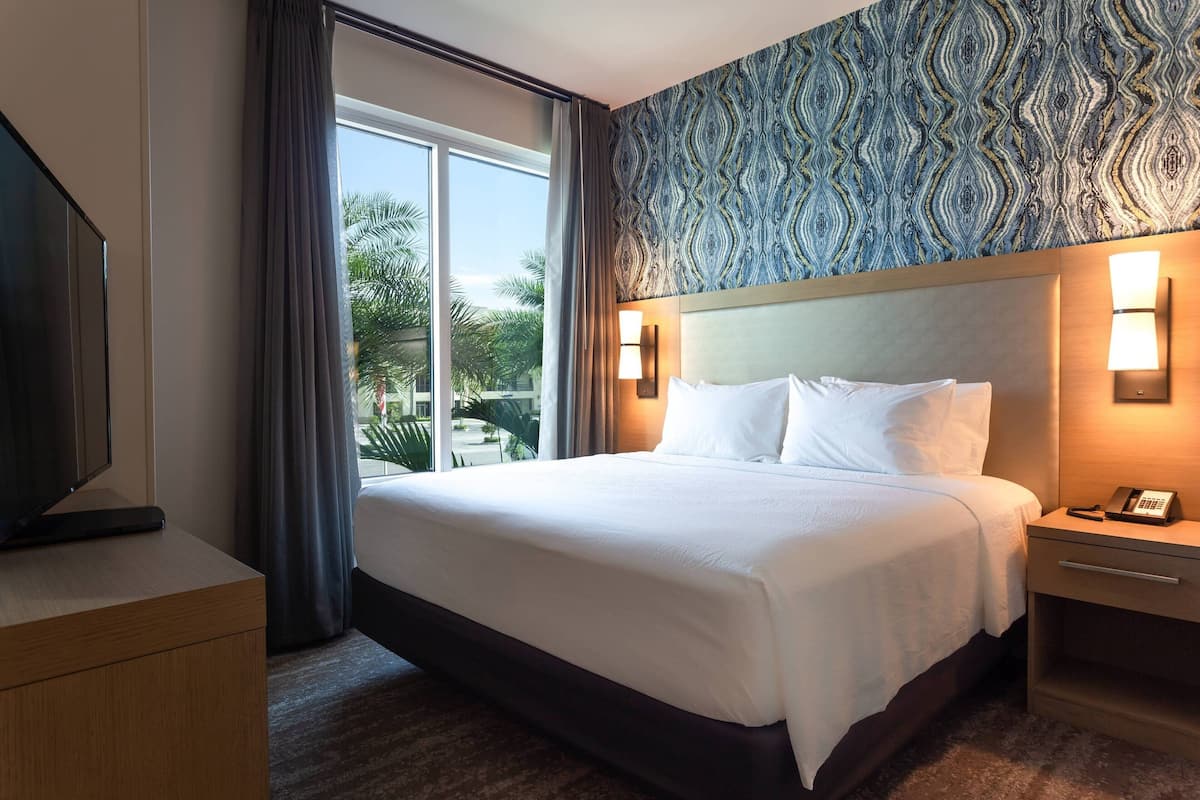 A hotel room with a large bed, a bedside table with a phone, a TV on a stand, and a window with curtains.