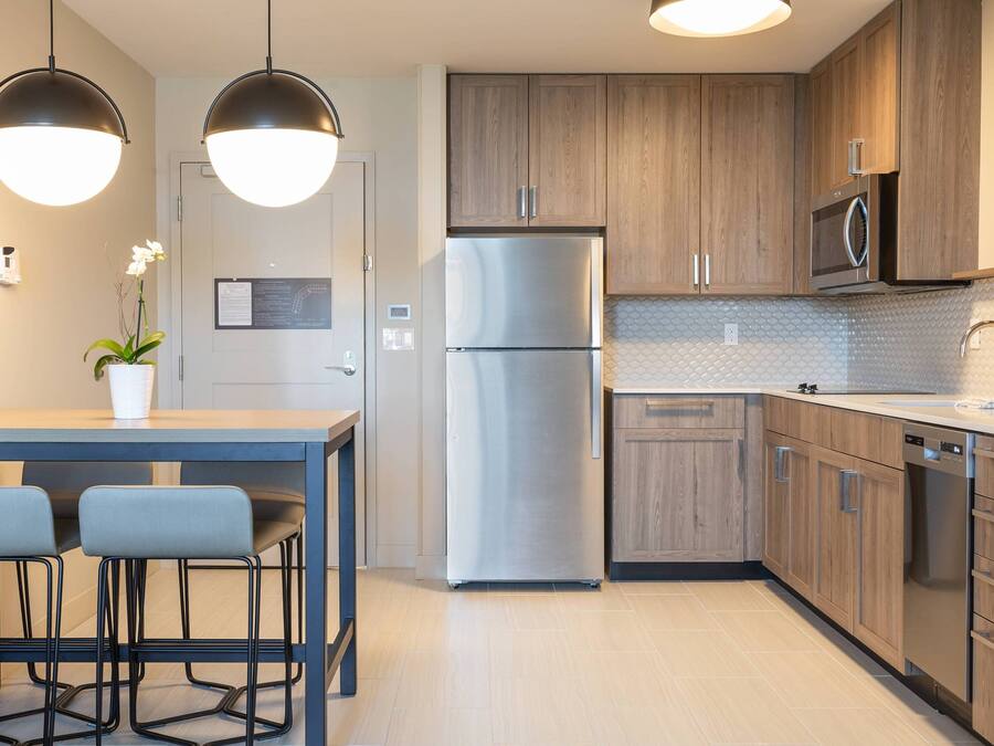Cocina moderna con armarios de madera, un refrigerador de acero inoxidable, un microondas y un lavavajillas. Hay una zona de comedor con una mesa y dos sillas.