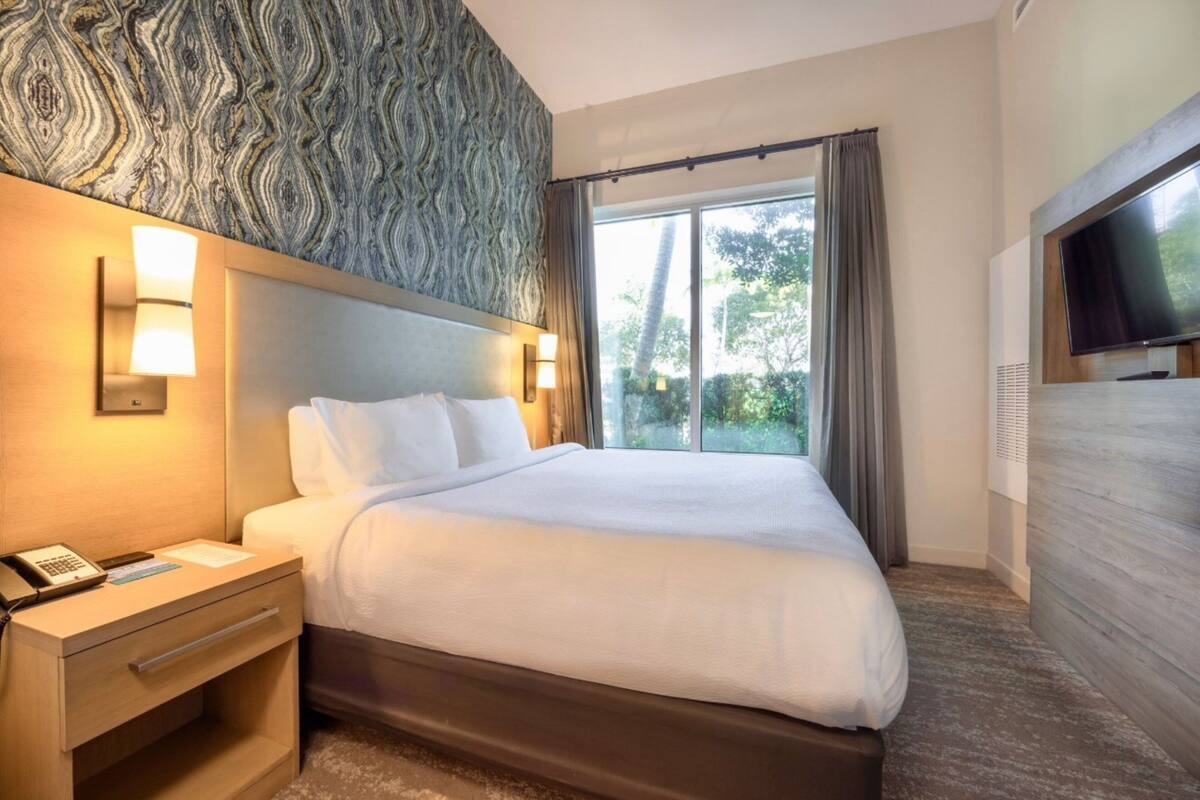 A hotel room with a large bed, bedside lamps, a flat-screen TV, and a view of the outdoors.