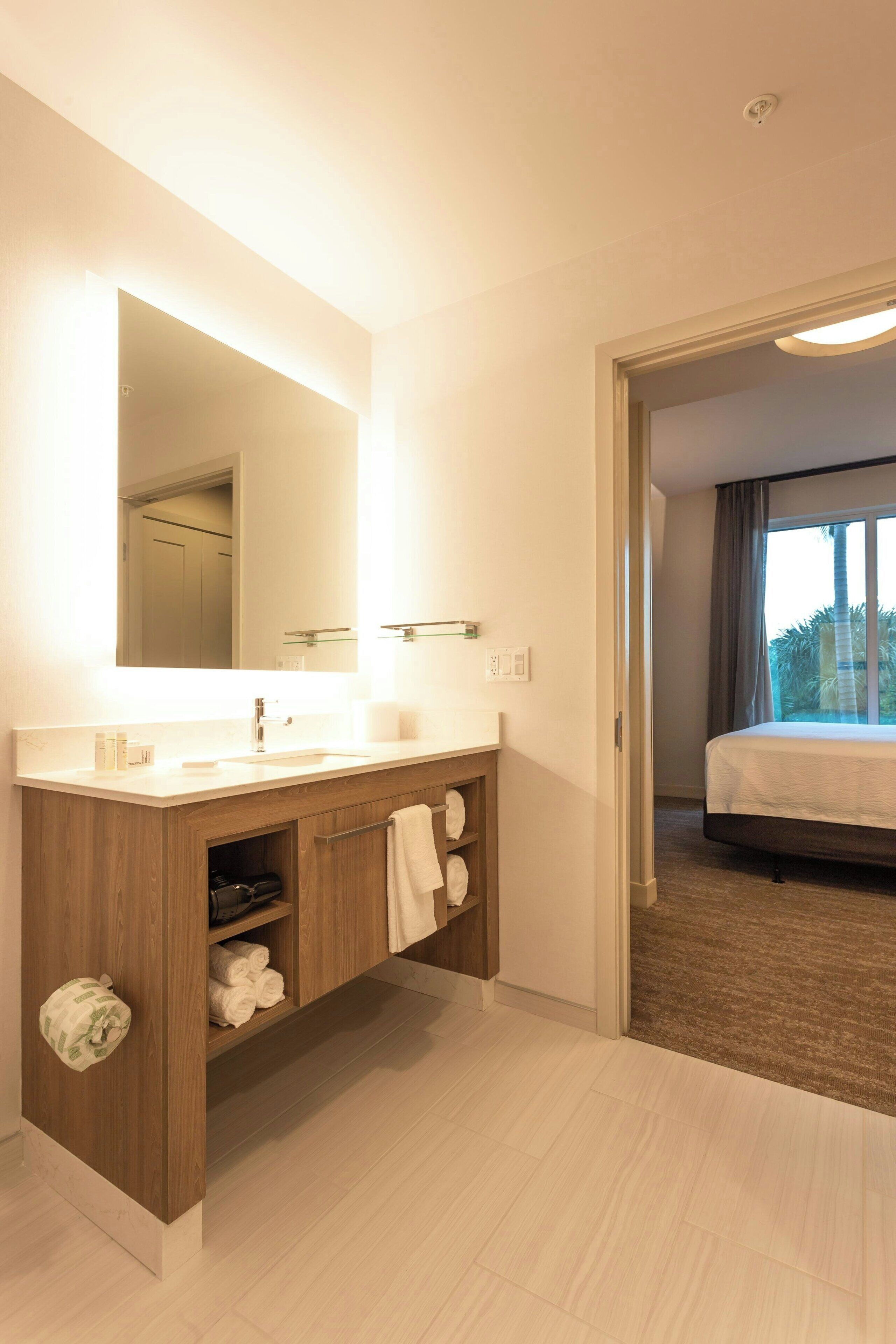 A modern bathroom with a large mirror, wooden vanity, and a view of a bedroom through an open door.