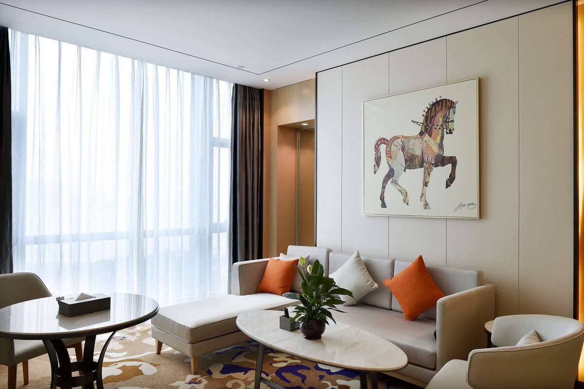 Une chambre d’hôtel moderne dotée d’une grande fenêtre, d’un ensemble de canapés, d’une table basse et d’un tableau représentant un cheval.