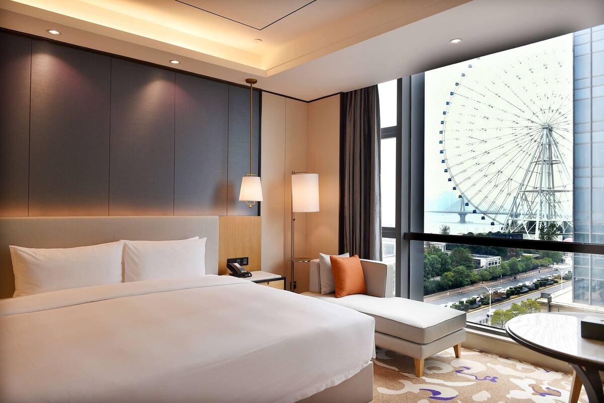 Une chambre d’hôtel avec un grand lit, un canapé, une petite table et une vue sur une grande roue, visible à travers une grande fenêtre.