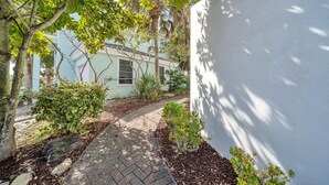 A paved pathway leading to a light blue building with a white door, surrounded by greenery and a white wall on one side. - January Beach Bliss for 2 OR 4!  Beach, Sunsets, pool, tennis!  US Senic 98 (Destin)