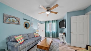 A living room with a sofa, coffee table, and a ceiling fan. There is a TV mounted on the wall and a door in the background. - January Beach Bliss for 2 OR 4!  Beach, Sunsets, pool, tennis!  US Senic 98 (Destin)