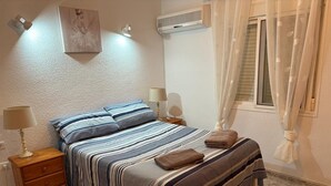 Une chambre à coucher comprenant un lit, des tables de chevet, une lampe, un tableau, un climatiseur et une fenêtre avec des rideaux.