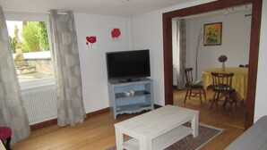 Ein Wohnzimmer mit Fernseher auf einem Schrank, einer weißen Bank und einem Essbereich mit Tisch und Stühlen.