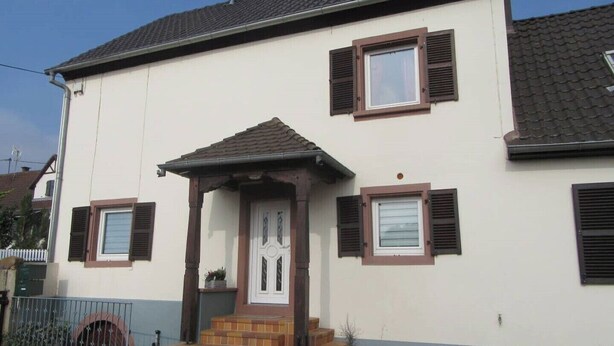 Ein zweistöckiges Haus mit weißer Fassade, braunen Fensterläden und einer kleinen Veranda mit Stufen.