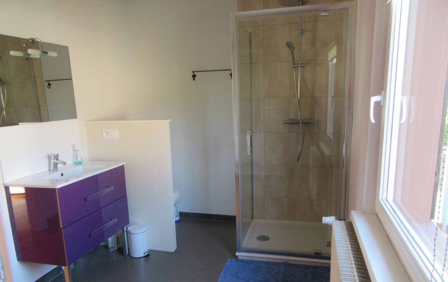A modern bathroom with a glass-enclosed shower, a purple vanity, and a window providing natural light.
