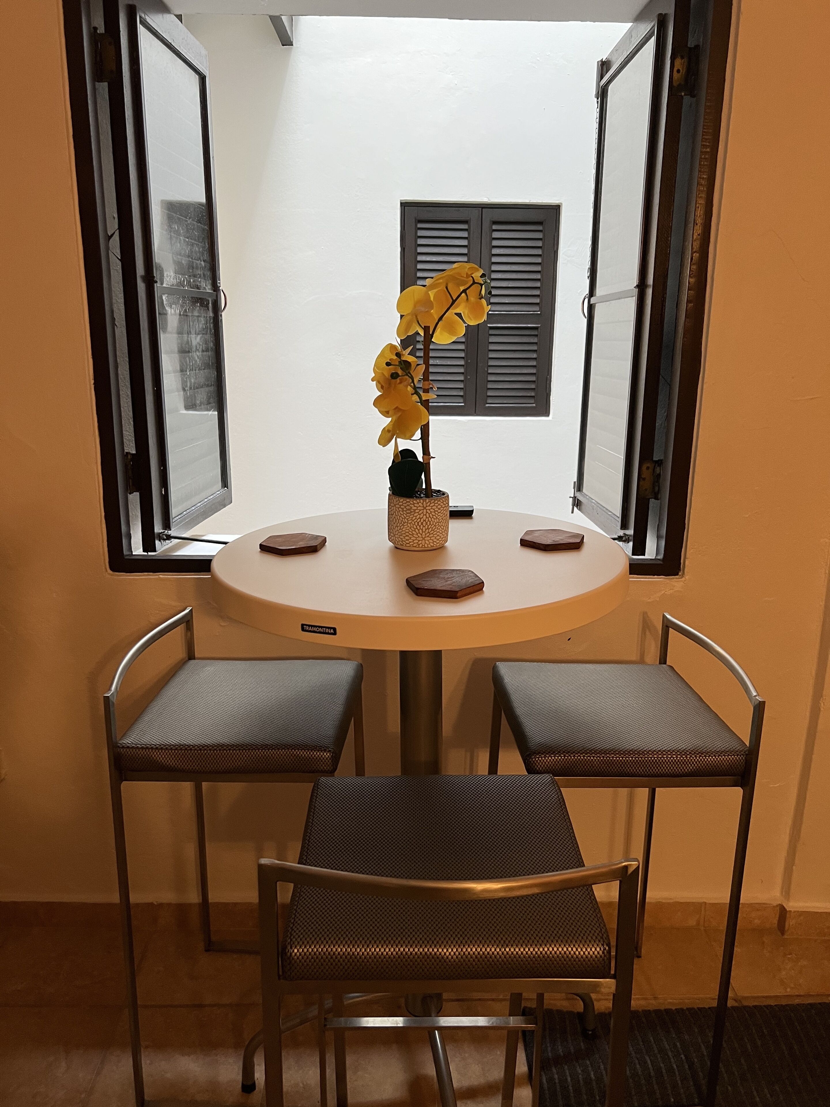 A round table with four chairs, a vase with yellow flowers, and a window with shutters.