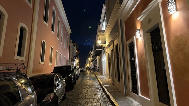 Une scène de rue nocturne avec un pavé, des voitures garées et des bâtiments dont les fenêtres et les portes sont illuminées.