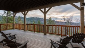 Una terraza techada con barandas de madera y sillas Adirondack, con vistas a un paisaje boscoso.