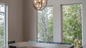 Un comedor con una gran mesa de madera y bancos, un colgante de luz y ventanas amplias con vista a árboles.