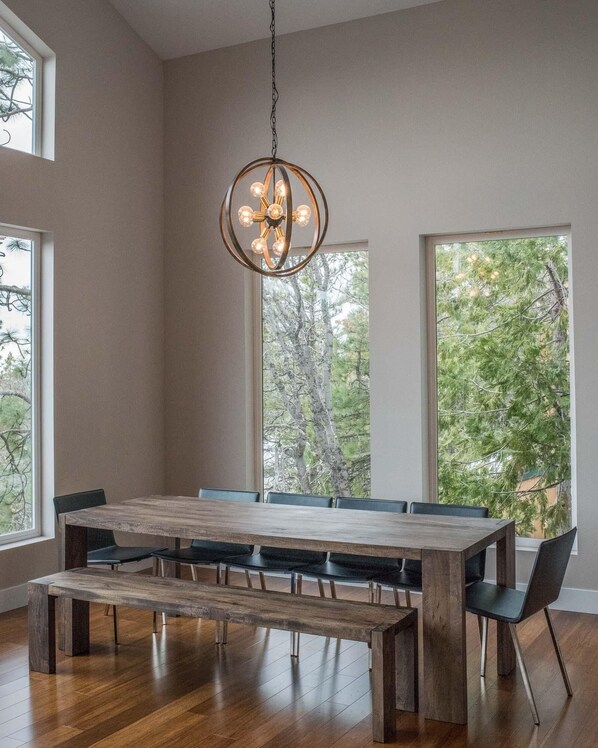 Un comedor con una gran mesa de madera y bancos, un colgante de luz y ventanas amplias con vista a árboles.