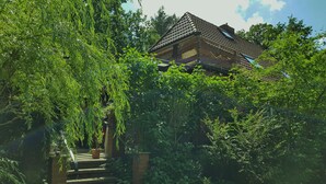 Ett traditionellt hus med ett tak täckt av tegelpannor, omgivet av frodig grönska och en trätrappa som leder upp till entrén.