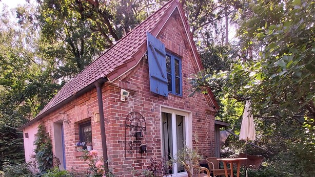 Ett rustikt hus med tegelmurar och ett sluttande tak, en blå träpersienn och en trädgård med krukväxter och blommor.