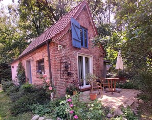 A rustic brick house with a sloped roof, a blue shutter, and a garden with potted plants and flowers. - Modernized stable with comfort and flair between forest and lake (Dobbertin)
