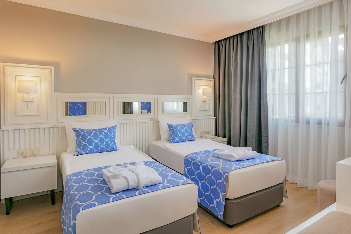 A hotel room with two beds, bedside tables, and a large window with curtains.