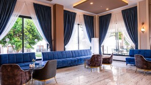 Eine moderne Hotellobby mit einem blauen Sofa, Sesseln, einem Glastisch und großen Fenstern mit Vorhängen.
