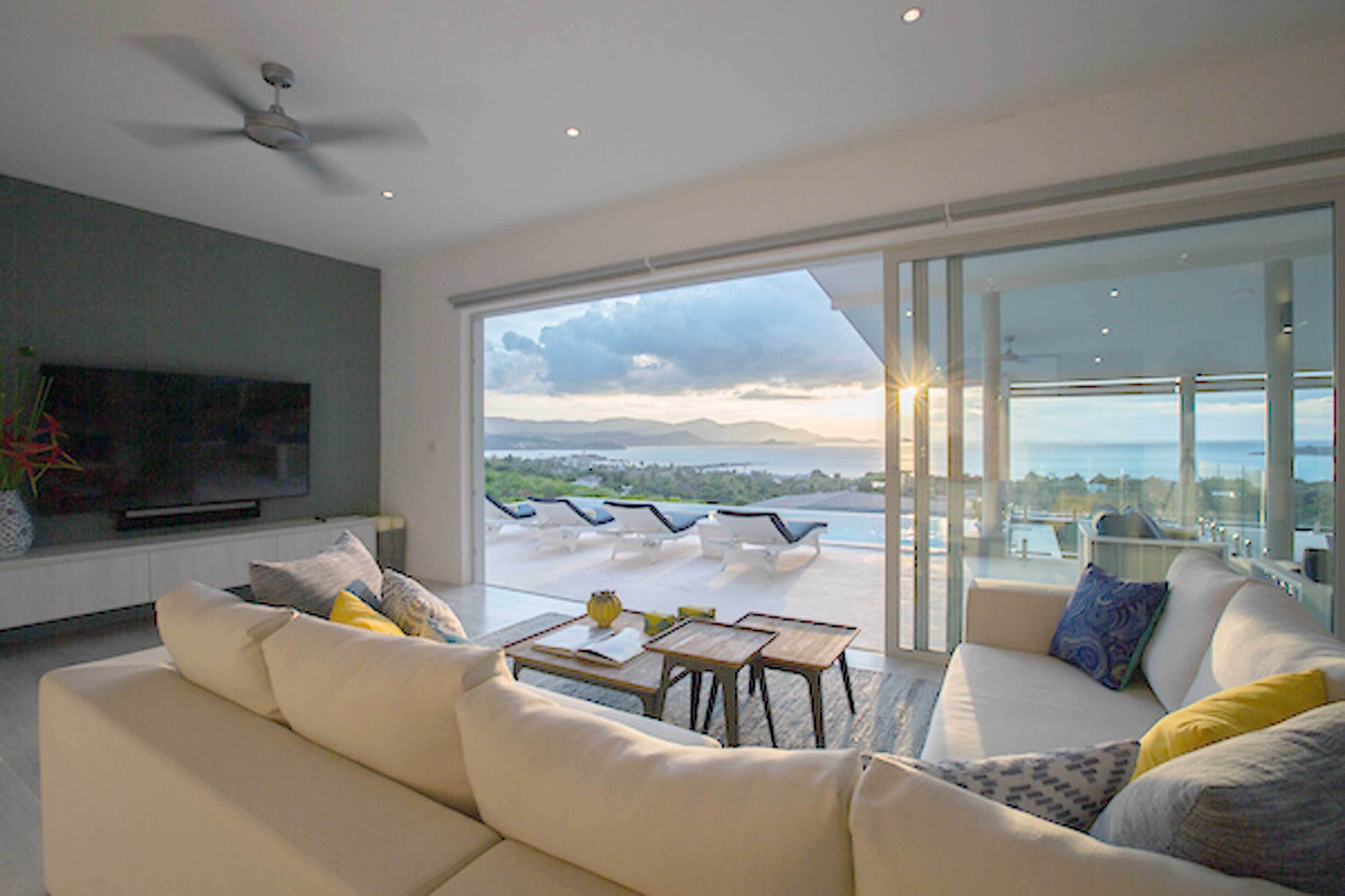 Una sala moderna con un televisor de pantalla plana grande, un sofá y una mesa de centro. Hay un balcón con asientos exteriores y vistas al paisaje.