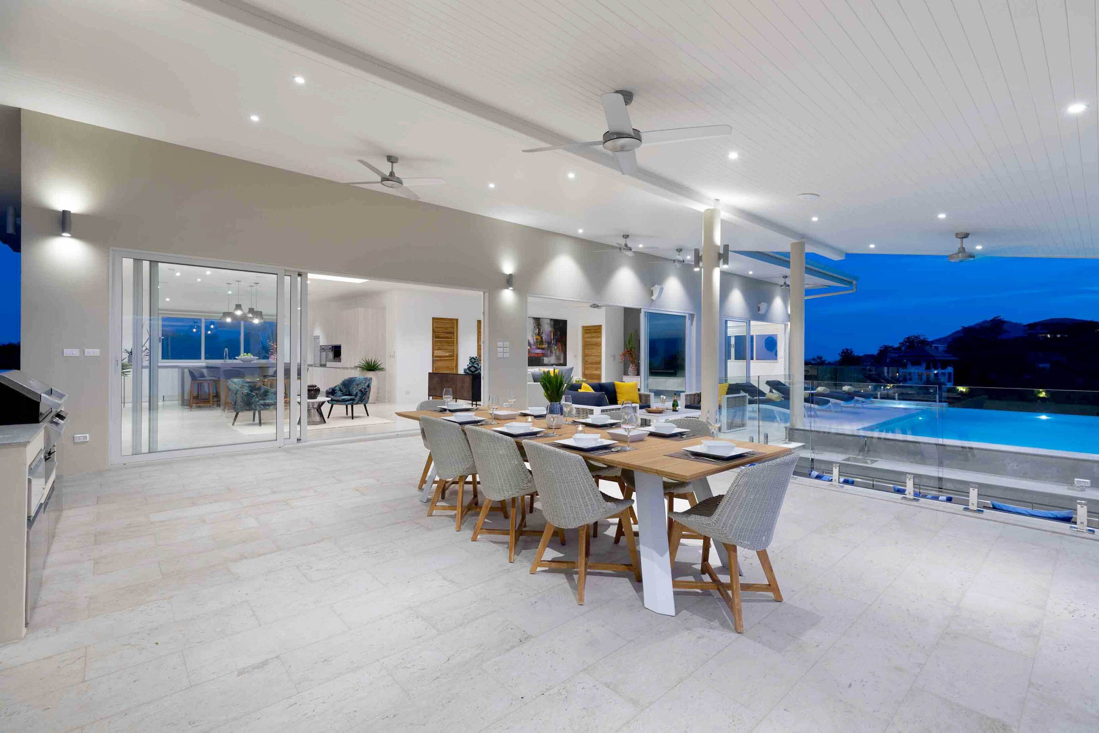 Un comedor moderno con vista a la piscina, que incluye una mesa larga con sillas, ventiladores de techo e iluminación empotrada.