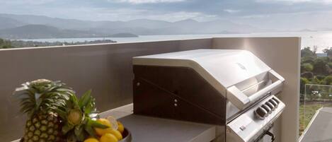 A balcony with a grill, a bowl of fruit, and a view of the sea and mountains.
