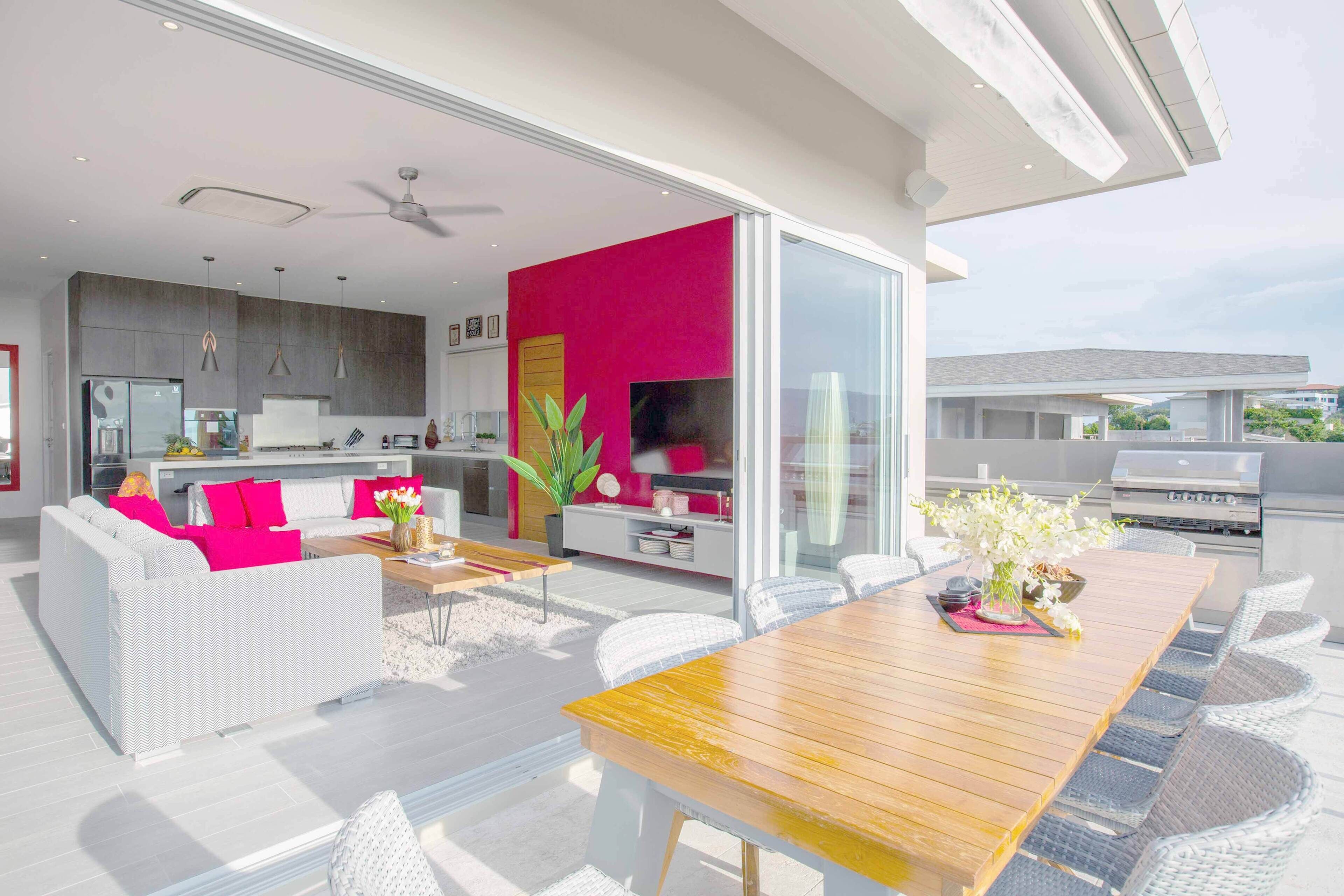 A modern kitchen with a large island, a dining area with a wooden table and white wicker chairs, and a bright pink accent wall.