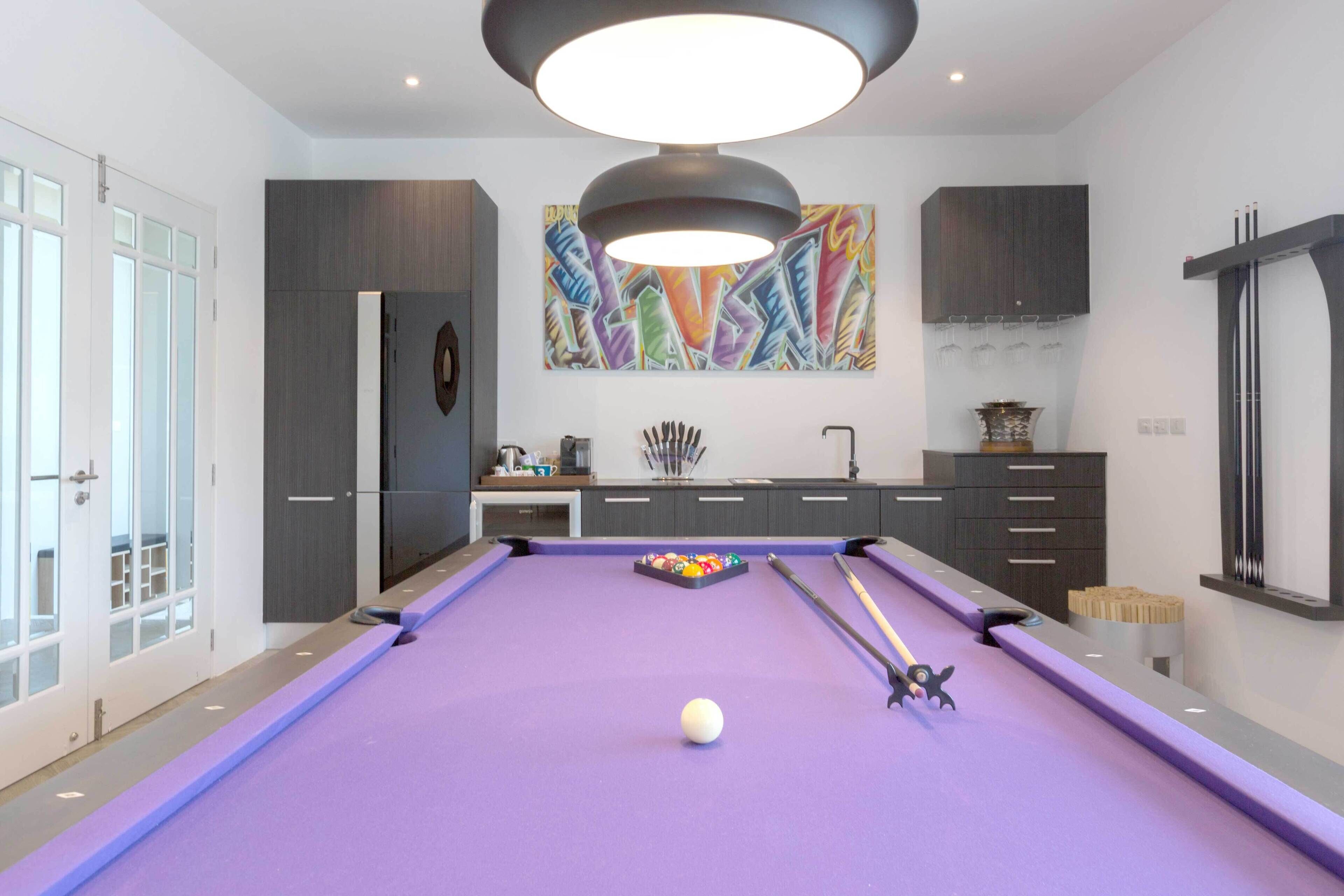 A modern pool table with a purple felt surface, a white cue ball, and pool cues.