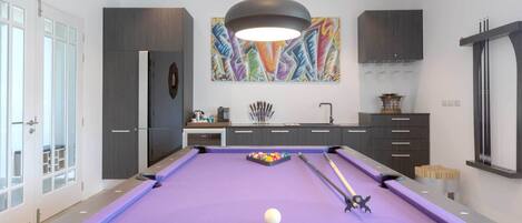 A modern pool table with a purple felt surface, a white cue ball, and pool cues.