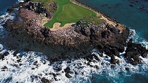 Vista aérea de un campo de golf costero con una isla rocosa en primer plano y una vista despejada del océano y la costa.