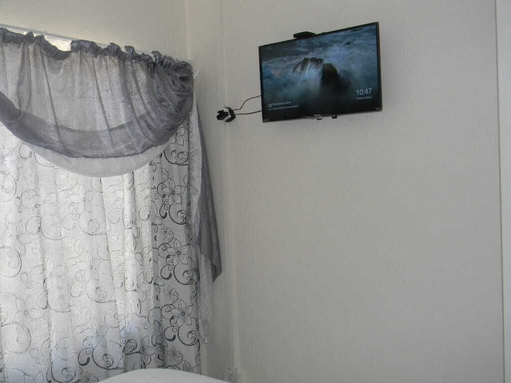 Een kamer met een bed, een aan de muur bevestigde televisie die een natuurlandschap toont, en een raam met dunne gordijnen.