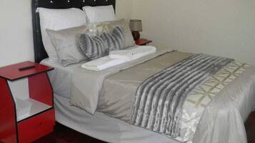 A neatly made bed with a grey and white duvet, a red nightstand with a lamp, and a wooden floor.
