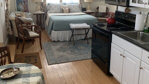 A compact, well-lit bedroom with a bed, bedside tables, and a window with curtains. Adjacent is a kitchenette with a stove, microwave, and cabinets. - Brookside Cottage is a beautiful open concept doll house stay for two. (Berkeley Springs)