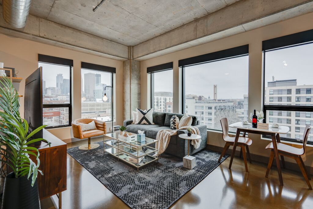 Una sala de estar moderna con sofá, mesa de centro y sillones, ventanas amplias con vista a la ciudad y una planta.