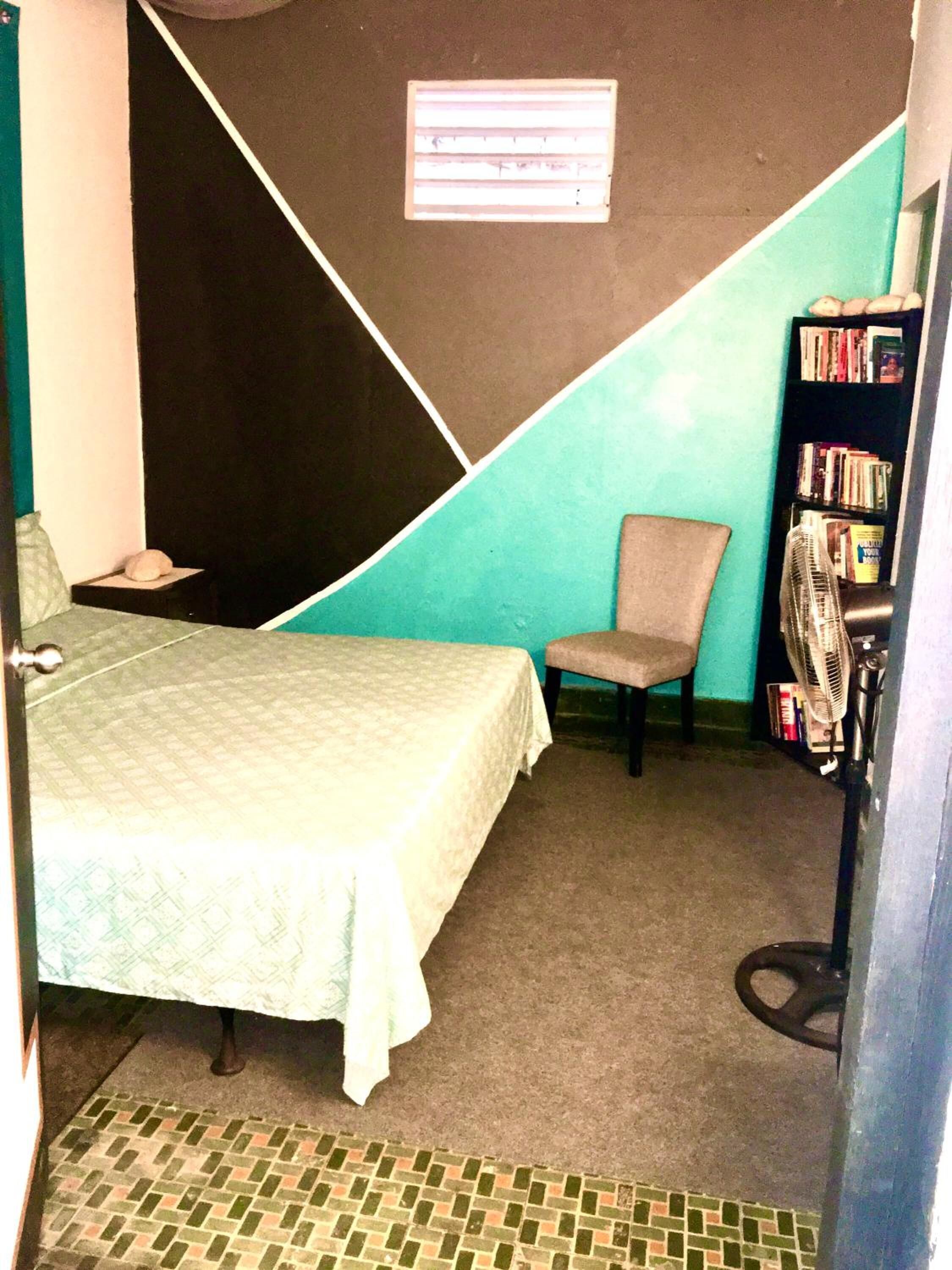 A small, compact room with a bed, a chair, a bookshelf, and a radiator.