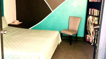 Una habitación pequeña y funcional con cama, sillón, estantería y radiador.