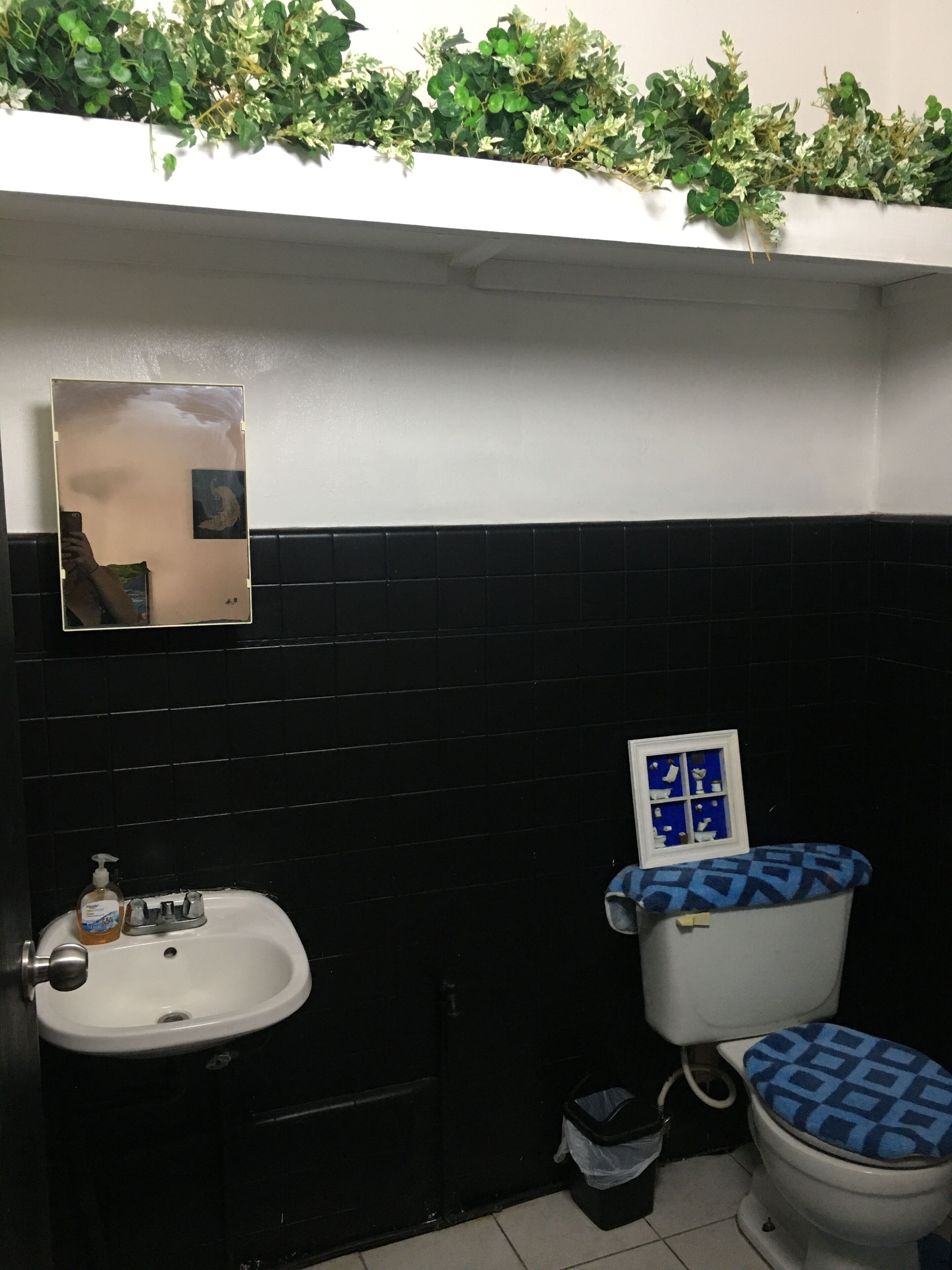 A bathroom with a white sink, black tiled walls, a toilet with a blue patterned seat cover, and a mirror with a picture frame.
