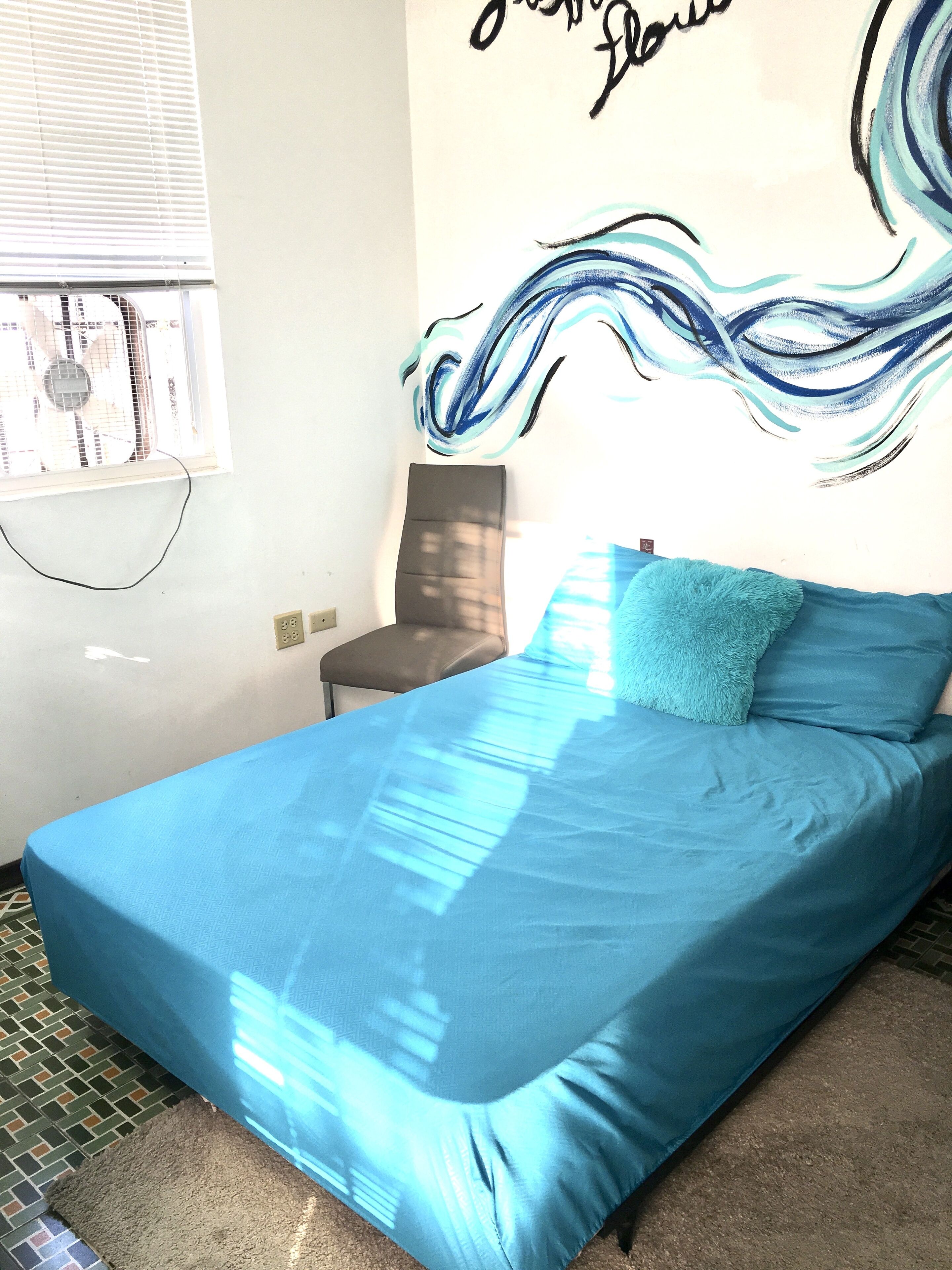 A bedroom with a blue bedspread, a grey chair, and a wall with a wave-like design.