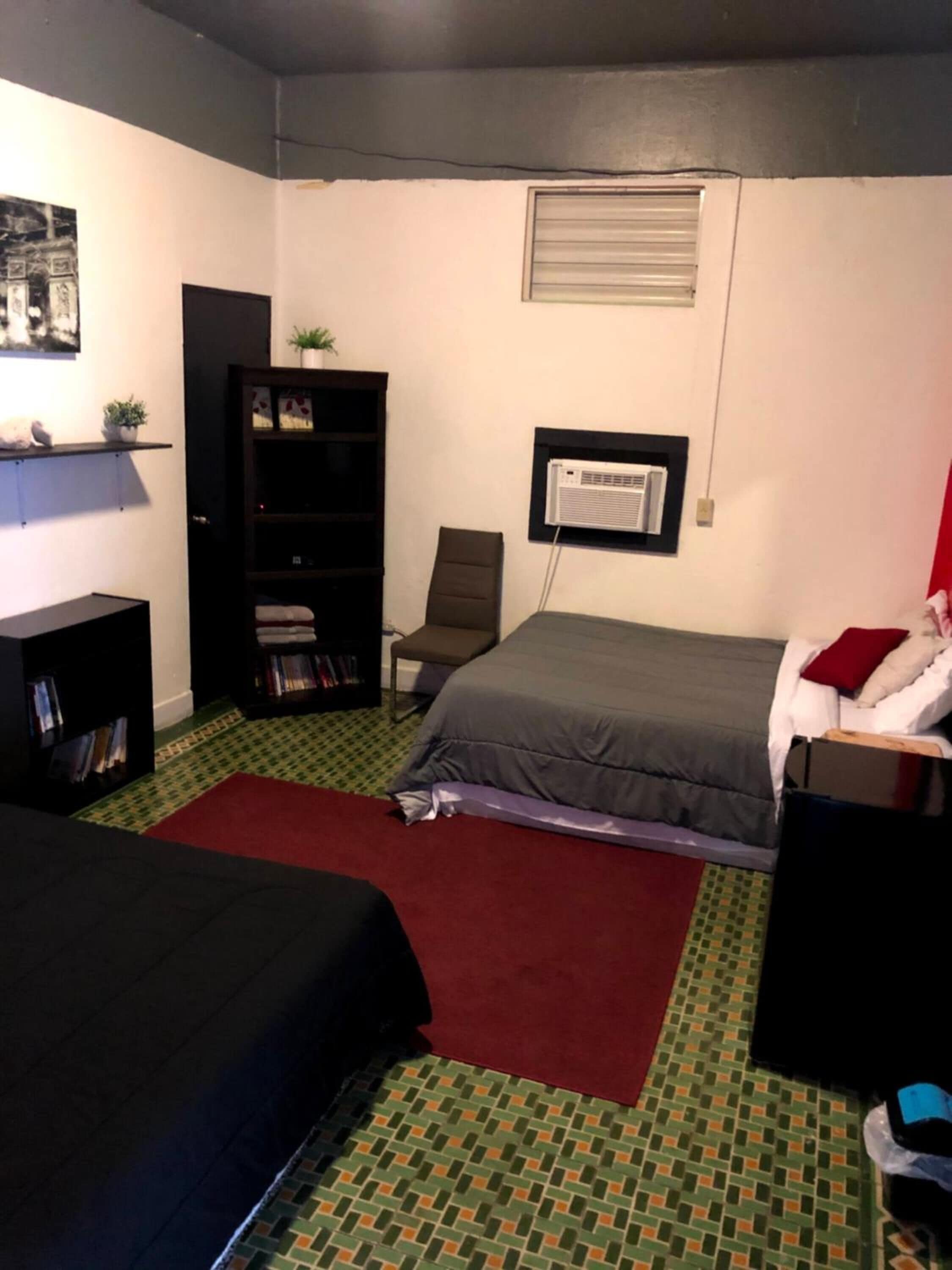 Ein Schlafzimmer mit zwei Betten, einem Bücherregal, einem Sessel und einem Fenster mit Klimaanlage.